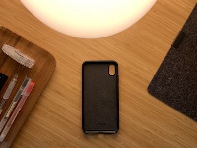 A wooden surface with a dark phone case, a glass desk lamp casting a warm glow, assorted pens and a clear tape dispenser on a wooden tray, and a portion of a dark gray felt item, possibly a mouse pad.