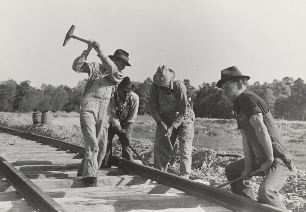 Fotografia em tons de cinza de quatro homens segurando martelo na ferrovia do trem