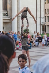 Two acrobats are performing a balancing act in front of an audience. One acrobat is standing and supporting her partner, who is upside down and arched backward in an impressive pose. The performance is taking place in an outdoor setting, possibly in a public square, with onlookers gathered around to watch.