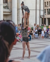 An acrobatic duo performing in a public square, wearing matching black sequined outfits. One performer is balanced on the shoulders of the other, surrounded by a crowd of seated and standing onlookers. The spectators appear engaged, with some taking photos.