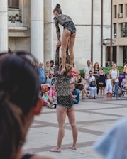 An acrobatic duo performing in a public square, wearing matching black sequined outfits. One performer is balanced on the shoulders of the other, surrounded by a crowd of seated and standing onlookers. The spectators appear engaged, with some taking photos.