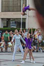 A young acrobat in a purple costume is being thrown into the air by two performers in matching outfits. The event takes place outdoors with a crowd watching, including a few seated people and some standing. The background features a modern building with large windows.