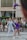 A young acrobat in a purple costume is being thrown into the air by two performers in matching outfits. The event takes place outdoors with a crowd watching, including a few seated people and some standing. The background features a modern building with large windows.