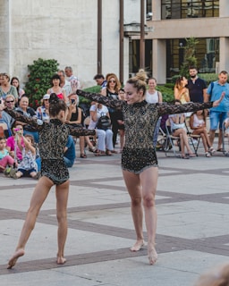 Two dancers perform in front of a seated audience outdoors, wearing matching black lace outfits. A diverse crowd of onlookers, including adults and children, watches attentively. The setting appears to be a public square with modern buildings in the background.