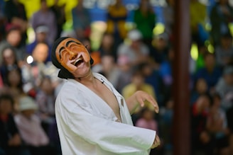 A person wearing a traditional mask is performing in front of a blurred crowd. The mask has an exaggerated expression of joy, and the person is dressed in a white garment. The audience in the background appears to be watching attentively.