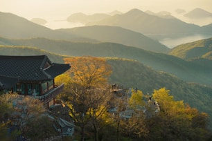 A traditional Asian temple is nestled among lush green hills, with a view of distant mountains and a serene body of water under a soft, golden light. The landscape features vibrant foliage and an expansive view of the layered terrain, creating a tranquil and picturesque scene.