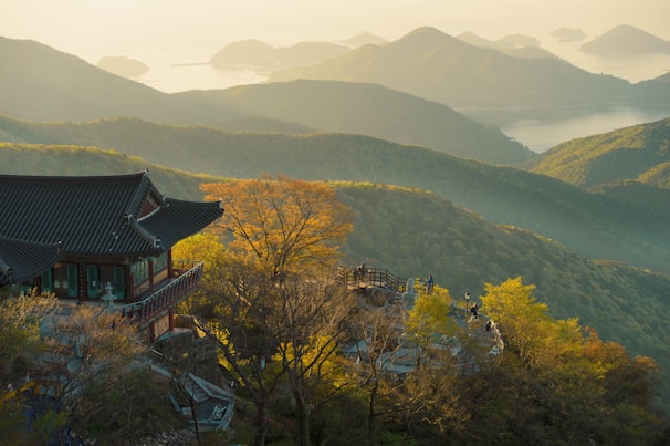 A traditional Asian temple is nestled among lush green hills, with a view of distant mountains and a serene body of water under a soft, golden light. The landscape features vibrant foliage and an expansive view of the layered terrain, creating a tranquil and picturesque scene.