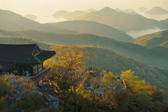 A traditional Asian temple is nestled among lush green hills, with a view of distant mountains and a serene body of water under a soft, golden light. The landscape features vibrant foliage and an expansive view of the layered terrain, creating a tranquil and picturesque scene.