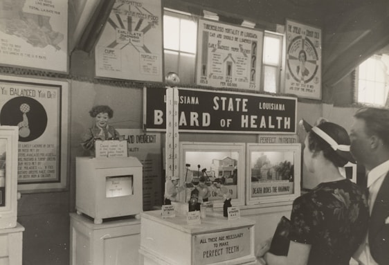 A black and white historical exhibit showcasing health-related information under the 'Louisiana State Board of Health' banner. Various posters and informational boards discuss topics such as tuberculosis, nutrition, and the importance of balanced diets. Two people are observing the displays closely, and there are several vintage medical and health-related artifacts visible.