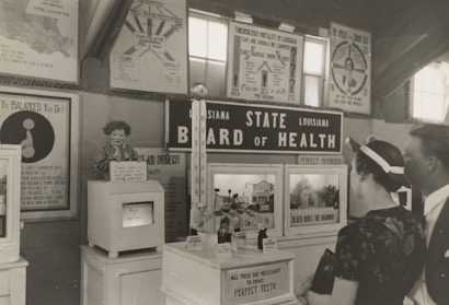 A black and white historical exhibit showcasing health-related information under the 'Louisiana State Board of Health' banner. Various posters and informational boards discuss topics such as tuberculosis, nutrition, and the importance of balanced diets. Two people are observing the displays closely, and there are several vintage medical and health-related artifacts visible.