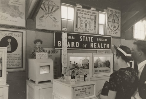 A black and white historical exhibit showcasing health-related information under the 'Louisiana State Board of Health' banner. Various posters and informational boards discuss topics such as tuberculosis, nutrition, and the importance of balanced diets. Two people are observing the displays closely, and there are several vintage medical and health-related artifacts visible.
