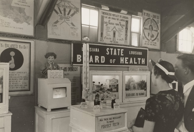 A black and white historical exhibit showcasing health-related information under the 'Louisiana State Board of Health' banner. Various posters and informational boards discuss topics such as tuberculosis, nutrition, and the importance of balanced diets. Two people are observing the displays closely, and there are several vintage medical and health-related artifacts visible.