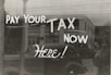 The image features a window display with bold, handwritten text reading 'PAY YOUR TAX NOW HERE!' A vintage car is visible in the reflective glass, along with a partial shadow or silhouette of a person. The background shows a street scene with a building.
