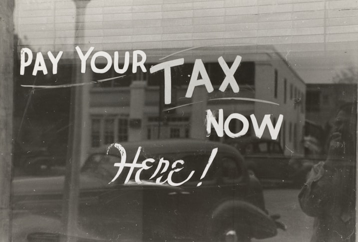The image features a window display with bold, handwritten text reading 'PAY YOUR TAX NOW HERE!' A vintage car is visible in the reflective glass, along with a partial shadow or silhouette of a person. The background shows a street scene with a building.
