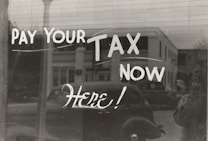 The image features a window display with bold, handwritten text reading 'PAY YOUR TAX NOW HERE!' A vintage car is visible in the reflective glass, along with a partial shadow or silhouette of a person. The background shows a street scene with a building.