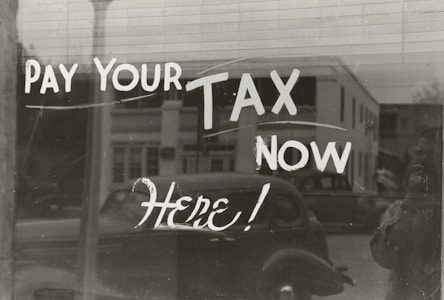 The image features a window display with bold, handwritten text reading 'PAY YOUR TAX NOW HERE!' A vintage car is visible in the reflective glass, along with a partial shadow or silhouette of a person. The background shows a street scene with a building.
