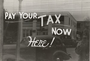 The image features a window display with bold, handwritten text reading 'PAY YOUR TAX NOW HERE!' A vintage car is visible in the reflective glass, along with a partial shadow or silhouette of a person. The background shows a street scene with a building.