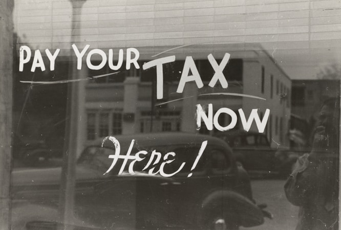 The image features a window display with bold, handwritten text reading 'PAY YOUR TAX NOW HERE!' A vintage car is visible in the reflective glass, along with a partial shadow or silhouette of a person. The background shows a street scene with a building.