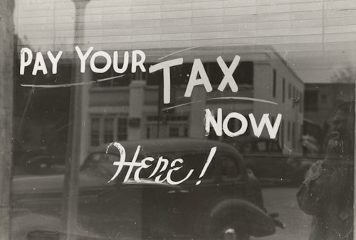 The image features a window display with bold, handwritten text reading 'PAY YOUR TAX NOW HERE!' A vintage car is visible in the reflective glass, along with a partial shadow or silhouette of a person. The background shows a street scene with a building.