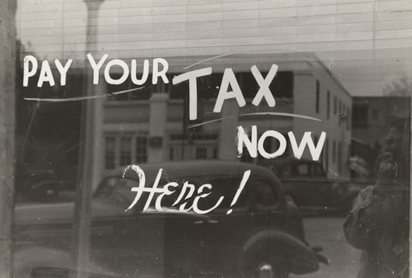 The image features a window display with bold, handwritten text reading 'PAY YOUR TAX NOW HERE!' A vintage car is visible in the reflective glass, along with a partial shadow or silhouette of a person. The background shows a street scene with a building.