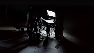 A dark and moody scene captures a film or photography set with crew members silhouetted against bright studio lights. They appear to be adjusting equipment and lighting setups, creating an atmosphere of focused activity. The scene is primarily lit by a large square panel light, illuminating parts of the set while casting deep shadows.