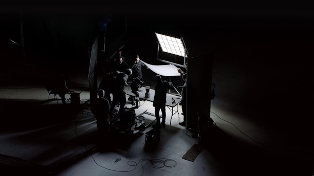 A dark and moody scene captures a film or photography set with crew members silhouetted against bright studio lights. They appear to be adjusting equipment and lighting setups, creating an atmosphere of focused activity. The scene is primarily lit by a large square panel light, illuminating parts of the set while casting deep shadows.