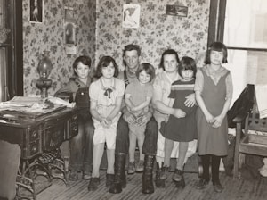 A family of six is sitting together in a room with floral wallpaper. The group consists of two adults and four children. The adults are seated on a bench while the children are gathered around them. On the left, an antique stove is covered with newspapers and a lamp. Decorative framed pictures hang on the walls, and the room appears to have a cozy, vintage atmosphere.