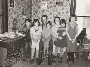 A family of six is sitting together in a room with floral wallpaper. The group consists of two adults and four children. The adults are seated on a bench while the children are gathered around them. On the left, an antique stove is covered with newspapers and a lamp. Decorative framed pictures hang on the walls, and the room appears to have a cozy, vintage atmosphere.