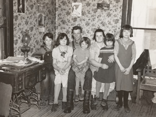 A family of six is sitting together in a room with floral wallpaper. The group consists of two adults and four children. The adults are seated on a bench while the children are gathered around them. On the left, an antique stove is covered with newspapers and a lamp. Decorative framed pictures hang on the walls, and the room appears to have a cozy, vintage atmosphere.
