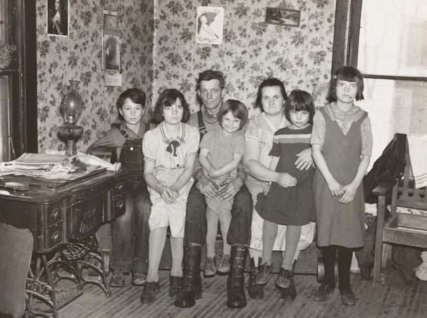 A family of six is sitting together in a room with floral wallpaper. The group consists of two adults and four children. The adults are seated on a bench while the children are gathered around them. On the left, an antique stove is covered with newspapers and a lamp. Decorative framed pictures hang on the walls, and the room appears to have a cozy, vintage atmosphere.