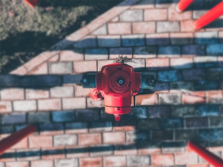 A red fire hydrant is situated on a paved surface with brick-like patterns. The pavement has alternating light and dark shades, and there is a small patch of grass visible at the top of the image.