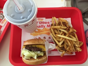 In a red tray, a meal is arranged with a cheeseburger wrapped in paper, a serving of fries, and a large soft drink cup with a straw. The background shows a fast-food restaurant logo.