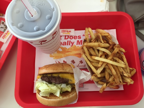 In a red tray, a meal is arranged with a cheeseburger wrapped in paper, a serving of fries, and a large soft drink cup with a straw. The background shows a fast-food restaurant logo.
