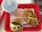 In a red tray, a meal is arranged with a cheeseburger wrapped in paper, a serving of fries, and a large soft drink cup with a straw. The background shows a fast-food restaurant logo.