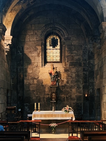 An ornate altar set against a stone wall featuring an arched window with geometric stained glass. A religious statue stands prominently on the altar, flanked by two lit candles and floral decorations. Warm lighting accentuates the texture of the stonework, creating a serene and contemplative atmosphere.
