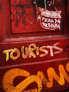 A red wall with various types of graffiti, including bold yellow spray-painted letters spelling 'TOURISTS' and additional abstract designs. A torn paper poster with faded text is also visible.