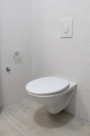 A modern, minimalistic bathroom featuring a wall-mounted toilet with a flush panel above it. The walls and floor are covered with light-colored tiles, and there is a toilet paper holder on the wall.