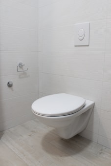 A modern, minimalistic bathroom featuring a wall-mounted toilet with a flush panel above it. The walls and floor are covered with light-colored tiles, and there is a toilet paper holder on the wall.