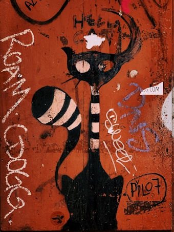 A mural on a wooden surface features a stylized black cat with large eyes and a long striped tail. The background is predominantly orange with various graffiti, text, and markings in different colors including white, purple, and black. The words 'HELLO CAT' are written above the cat, and the word 'SWEET' is written beside it.