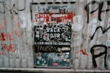 A metal door on a corrugated wall is covered with various stickers, graffiti, and posters advertising live music shows. The text includes phrases like 'EVERY FRIDAY 10PM' and 'BACK DOOR'. The surrounding wall also features graffiti in different colors.