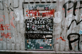 A metal door on a corrugated wall is covered with various stickers, graffiti, and posters advertising live music shows. The text includes phrases like 'EVERY FRIDAY 10PM' and 'BACK DOOR'. The surrounding wall also features graffiti in different colors.