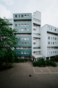 A multi-story residential building with a modern design, featuring teal and white exterior walls. The structure includes numerous windows and a slightly recessed entrance with a small overhang. In front of the building, there is a designated parking space for people with disabilities, indicated by yellow markings on the pavement. Lush greenery and a large tree are present on the left side, adding a natural element to the urban setting. The overall scene appears quiet and empty, with no visible people or vehicles.