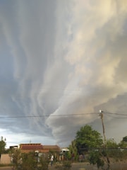 Dramatic, swirling storm clouds fill the sky above a small rural area with houses and trees. Power lines stretch across the scene, and a few people are visible on the ground.