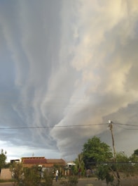 Dramatic, swirling storm clouds fill the sky above a small rural area with houses and trees. Power lines stretch across the scene, and a few people are visible on the ground.