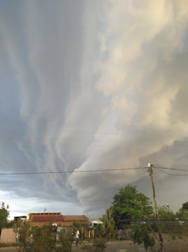 Dramatic, swirling storm clouds fill the sky above a small rural area with houses and trees. Power lines stretch across the scene, and a few people are visible on the ground.