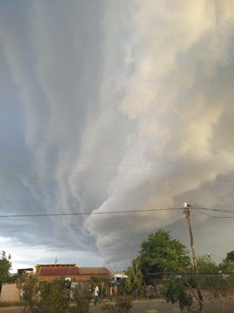 Dramatic, swirling storm clouds fill the sky above a small rural area with houses and trees. Power lines stretch across the scene, and a few people are visible on the ground.