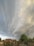 Dramatic, swirling storm clouds fill the sky above a small rural area with houses and trees. Power lines stretch across the scene, and a few people are visible on the ground.