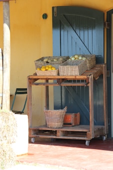 A rustic wooden cart holds several wicker baskets filled with a variety of vegetables and fruits, such as lemons and potatoes. The cart is placed against a textured, mustard-yellow wall with a dark green door, creating a cozy ambiance. There is also a small, classic green chair nearby and a stack of hay to the left.