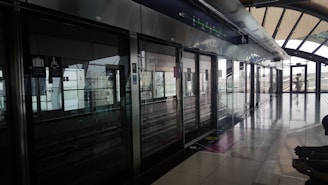 A modern, clean station platform with sleek metallic finishing and large glass windows. Signs for luggage and accessibility features are visible above sliding doors. The platform is empty, emphasizing a quiet and orderly environment with natural light streaming in.