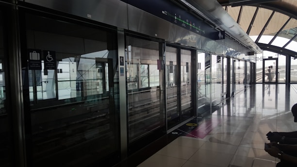 A modern, clean station platform with sleek metallic finishing and large glass windows. Signs for luggage and accessibility features are visible above sliding doors. The platform is empty, emphasizing a quiet and orderly environment with natural light streaming in.